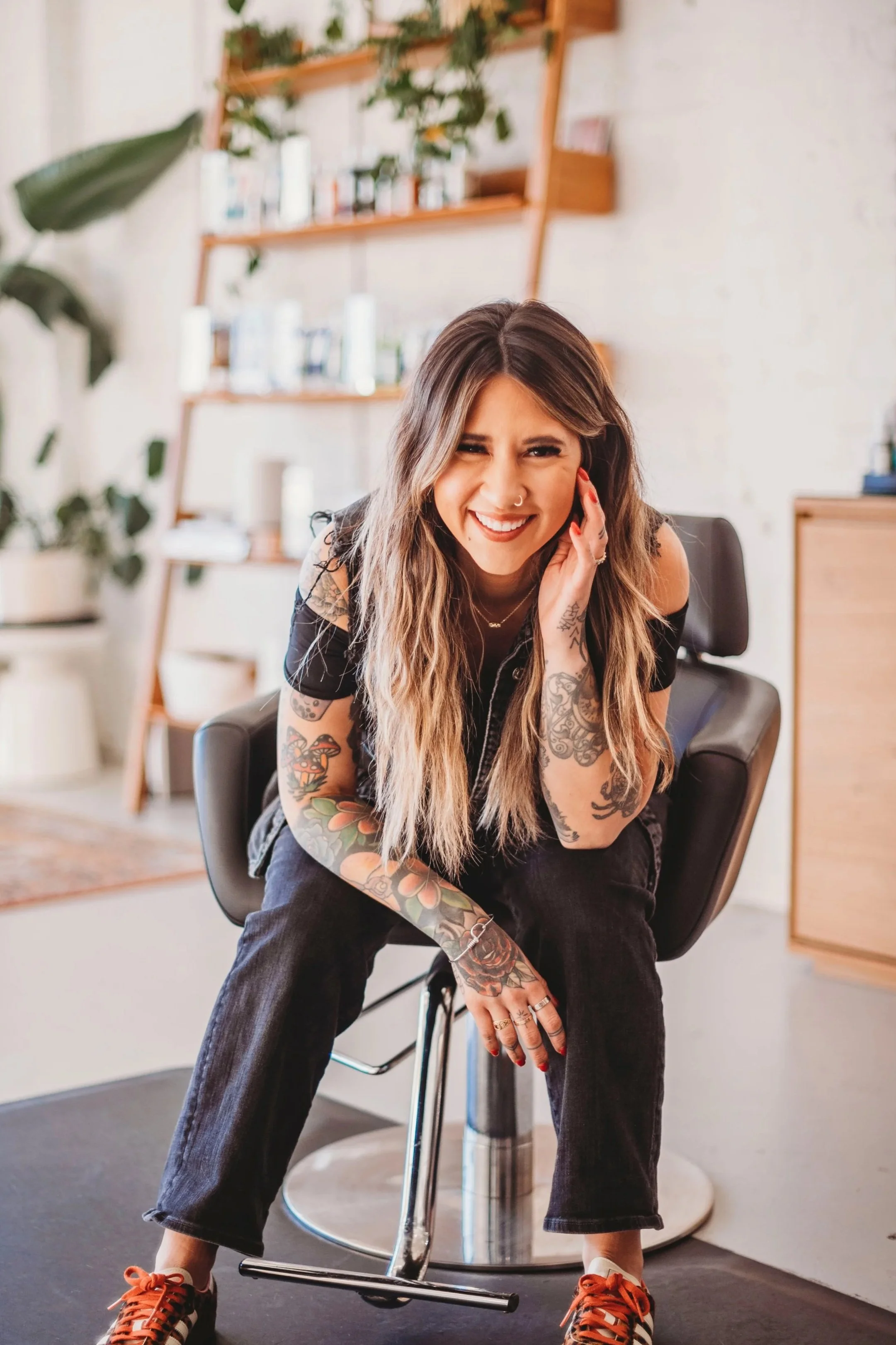 A woman with long, wavy brown hair and multiple colorful tattoos on her arms, wearing orange sneakers and black pants, sitting on a black chair in a modern room with wooden shelves, houseplants, and a light-colored wall in the background.