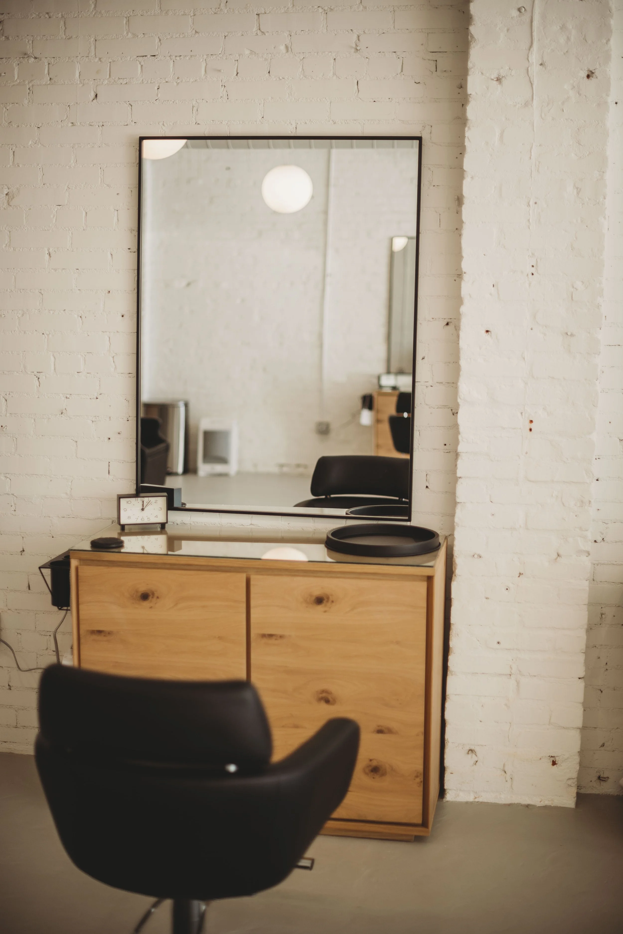 A modern salon setup featuring a wooden counter with a large rectangular mirror above it, a black salon chair in front, a small digital clock on the counter, and a black tray with styling tools, all against a white brick wall.