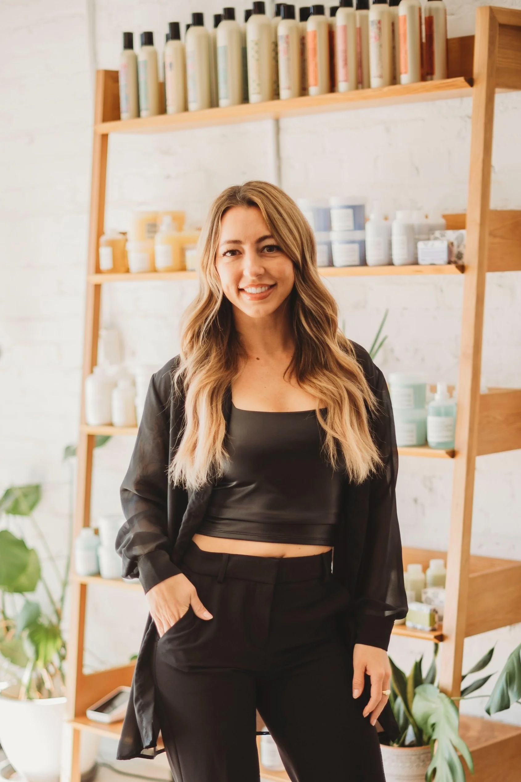 A woman with long wavy hair standing in front of wooden shelves filled with bottles and containers, smiling and wearing a black satin crop top and matching pants.