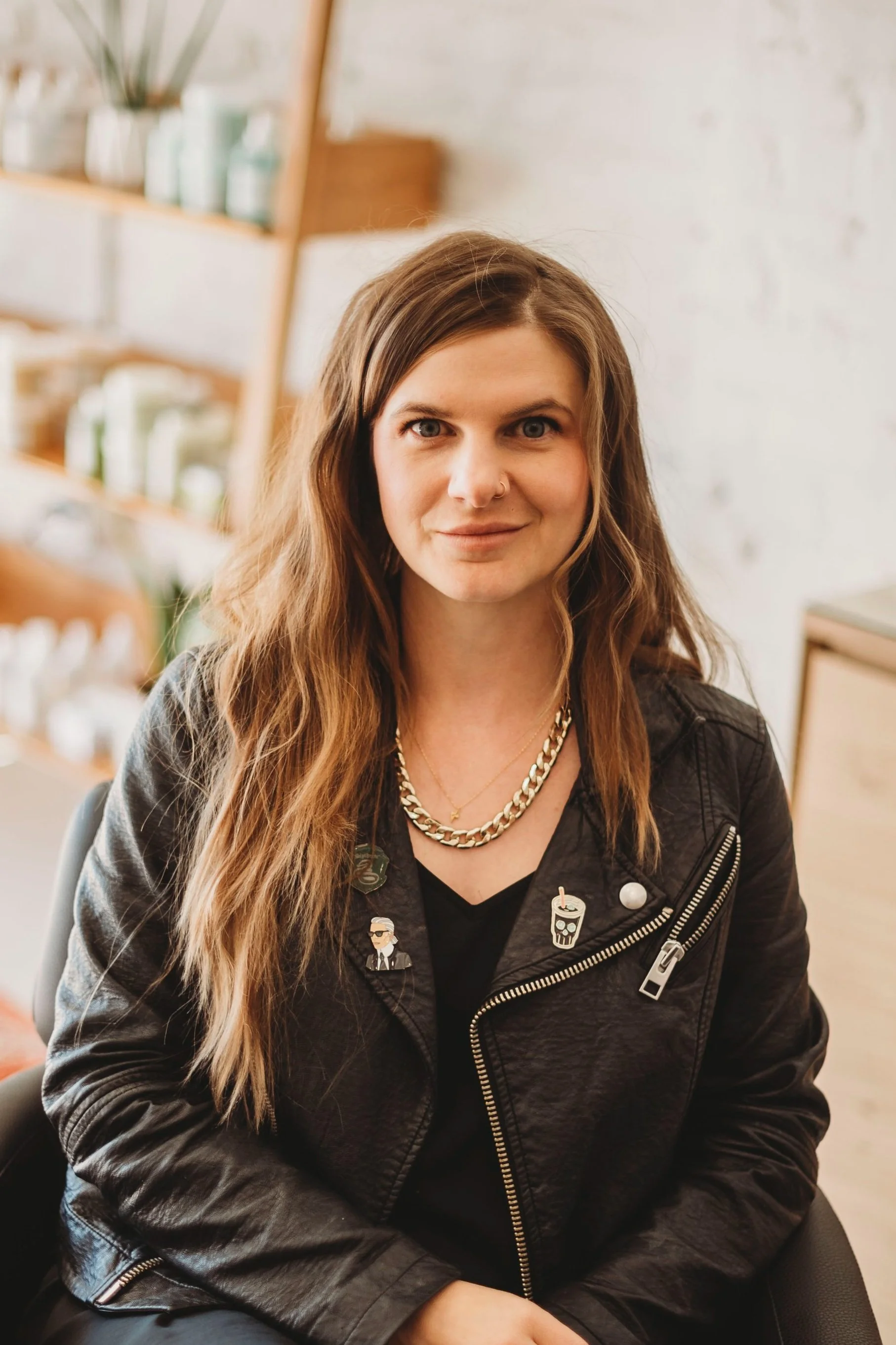 A woman with long wavy brown hair, wearing a black leather jacket with pins, a gold chain necklace, and a black top, sitting in a room with wooden shelves and plants in the background.