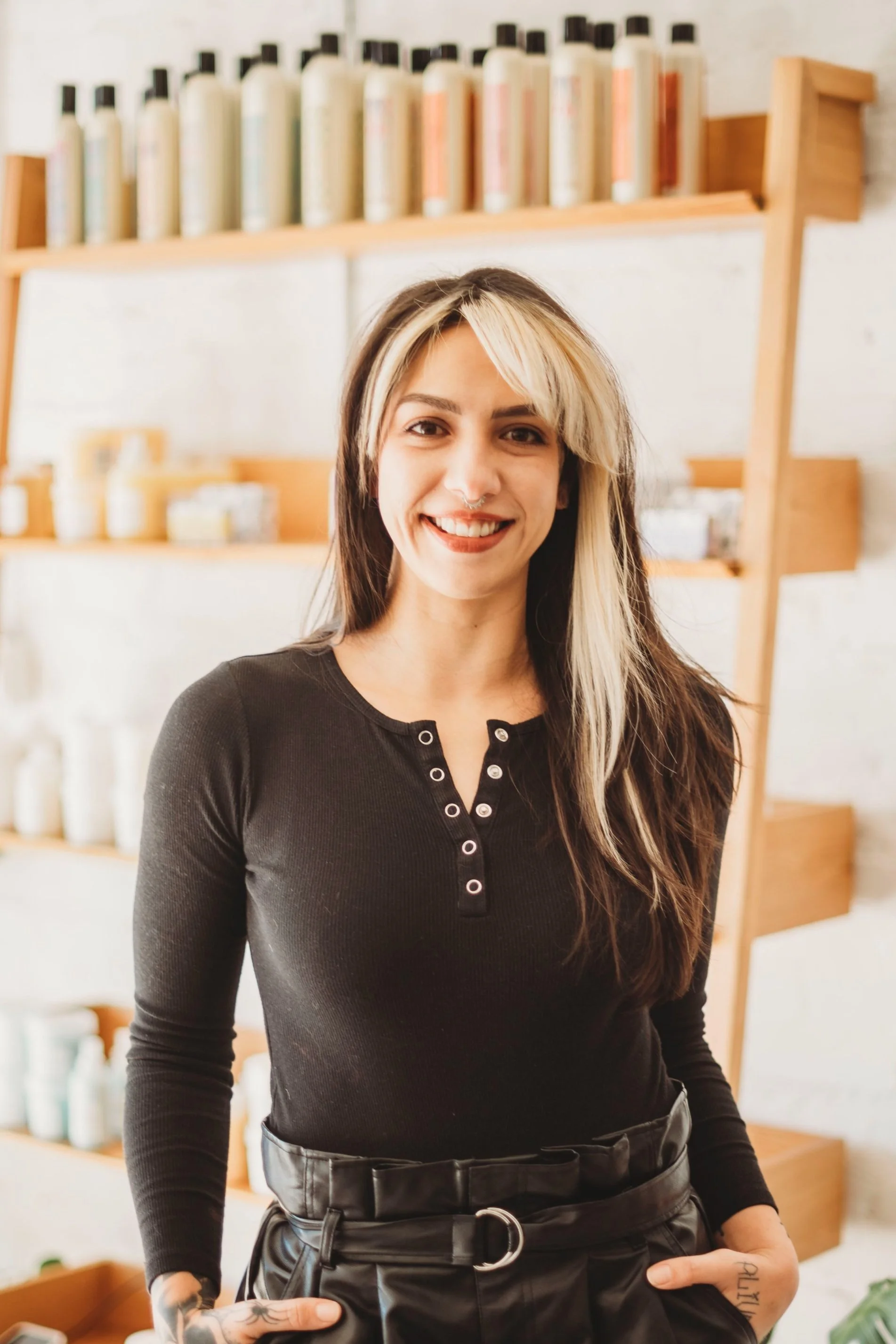 A smiling woman with blonde and brown hair, wearing a black long-sleeve shirt and black leather pants, standing in front of wooden shelves with bottles of skincare or haircare products.
