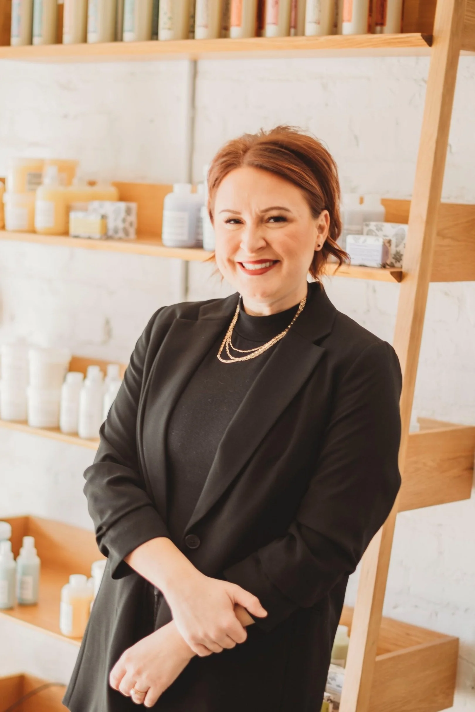 A woman with red hair wearing a black blazer and necklaces, smiling and standing in front of wooden shelves with bottles and boxes in a retail or pharmacy setting.