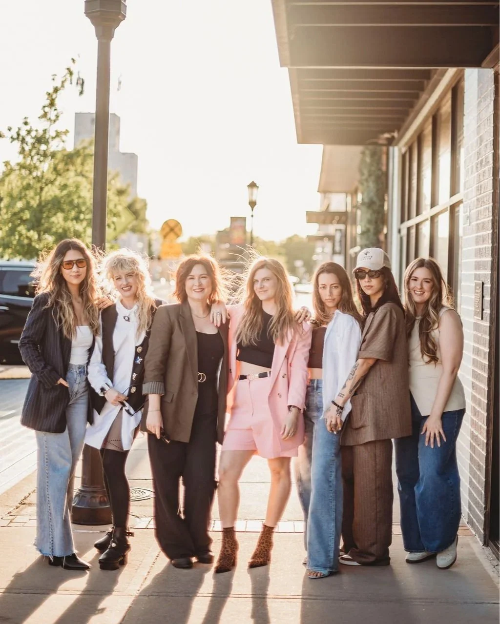 Group of seven women standing on sidewalk outdoors during sunset, smiling, in casual and stylish clothing.